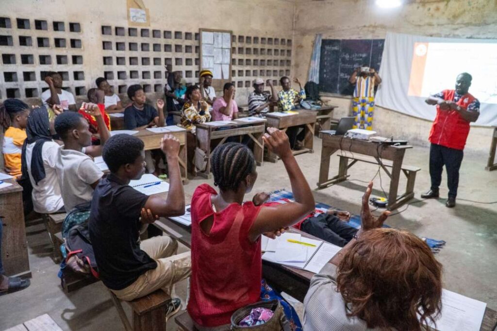 Capacity building in disaster management, A classroom setting where a group of people, including youth and adults, are engaged in a training session led by a presenter in a red vest. The participants are seated at wooden desks in a simple room, with some raising their hands to answer or participate. A projector screen is displayed at the front with information, suggesting a capacity-building training in disaster management. The atmosphere is interactive and focused on learning and preparedness.